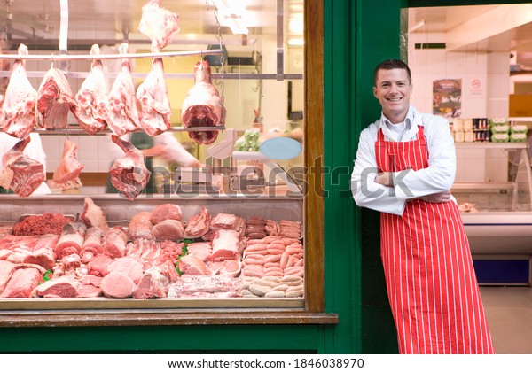 A young smiling\
butcher in red apron leaning against butcher shop doorway next to\
the display window.