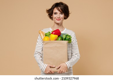 Young smiling caucasian cheerful happy fun vegetarian woman 20s in casual clothes hold paper bag with vegetables after shopping look camera isolated on plain pastel beige background studio portrait