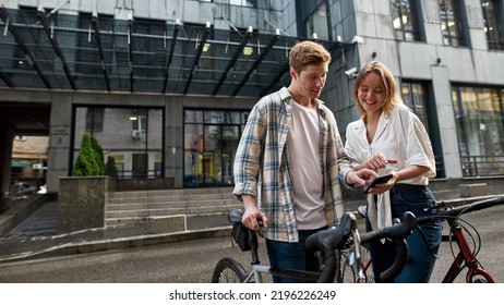 Young smiling caucasian couple with bicycles watching and pointing at smartphone on city street. Modern urban lifestyle. Entertainment, leisure and hobby. Biking. Man and girl spending time together - Powered by Shutterstock