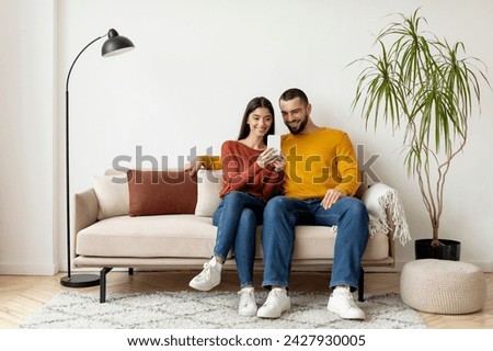 Young Smiling Caucasian Couple Using Mobile Phone And Embracing Together At Home, Happy Millennial Spouses Relaxing On Couch With Modern Smartphone, Browsing New App Or Shopping Online, Copy Space