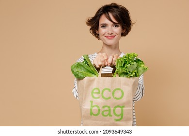 Young smiling caucasian european happy fun vegetarian woman 20s in casual clothes hold paper bag with vegetables after shopping look camera isolated on plain pastel beige background studio portrait