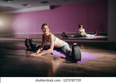 Young smiling Caucasian fit brunette sitting on the mat and stretching legs while wearing kangoo jumps boots. Dark gym interior.