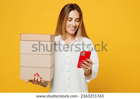 Young smiling caucasian happy woman she wears white shirt casual clothes hold stack cardboard blank boxes use mobile cell phone isolated on plain yellow background studio portrait. Lifestyle concept