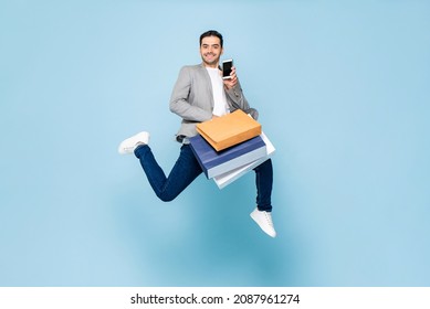 Young Smiling Caucasian Man Holding Shopping Bags And Mobile Phone While Jumping In Light Blue Isolated Background