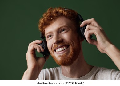 Young Smiling Caucasian Red Hair Guy Taking Off Earphones From Ears And Looking Away. Concept Of Human Of Generation Z. Handsome Bearded Male Person. Isolated On Dark Green Background In Studio