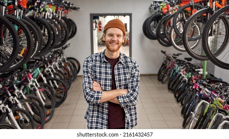 Young smiling caucasian redhead male seller with crossed arms posing and looking at camera in modern bike shop. Set of variety bikes for extreme sport, urban lifestyle and children. Shopping concept