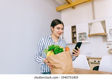 Young Smiling Caucasian Woman Use Smartphone In The Modern Kitchen, Bag With Fresh Vegetable On The Table. Online Buying Food And Grocery.