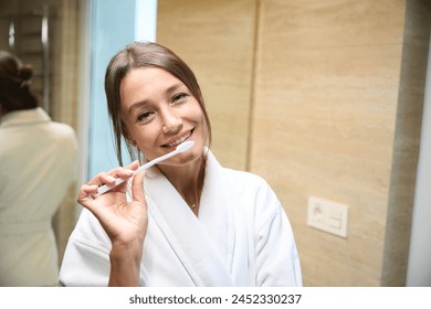 Young smiling caucasian woman wearing bathrobe brushing teeth with toothbrush - Powered by Shutterstock