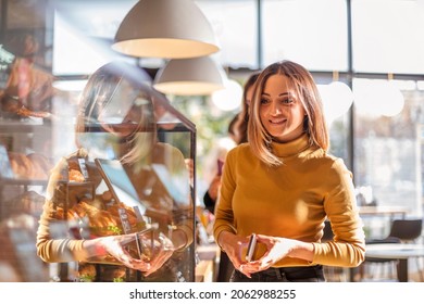 Young smiling charming woman standing in line at cafe or coffee shop on morning, hungry female with mobile phone in hands choosing food while standing near showcase at bakery shop - Powered by Shutterstock