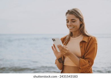 Young smiling cheerful Caucasian woman she wearing orange shirt casual clothes hold in hand use mobile cell phone walk on sea ocean sand shore beach outdoor seaside in summer day. Lifestyle concept - Powered by Shutterstock