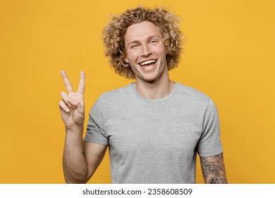 Young smiling cheerful fun cool positive joyful caucasian man 20s he wear grey t-shirt look camera showing victory sign isolated on plain yellow background studio portrait. People lifestyle concept.