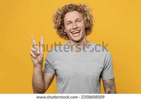Young smiling cheerful fun cool positive joyful caucasian man 20s he wear grey t-shirt look camera showing victory sign isolated on plain yellow background studio portrait. People lifestyle concept.
