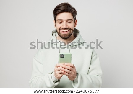 Young smiling cheerful happy caucasian man wear mint hoody hold in hand use mobile cell phone in green case chatting isolated on plain solid white background studio portrait. People lifestyle concept