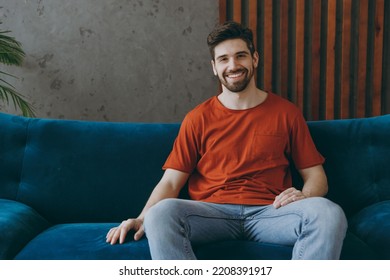 Young smiling cheerful happy fun european man wears red t-shirt sit on blue sofa couch stay at home hotel flat rest relax spend free spare time in living room indoors grey wall. People lounge concept - Shutterstock ID 2208391917