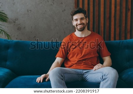 Young smiling cheerful happy fun european man wears red t-shirt sit on blue sofa couch stay at home hotel flat rest relax spend free spare time in living room indoors grey wall. People lounge concept