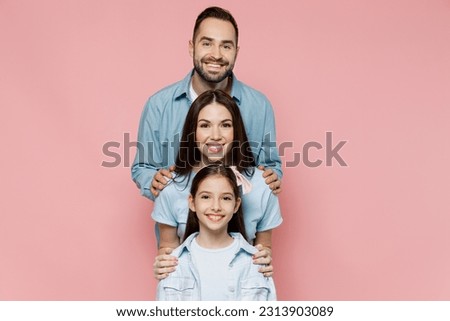 Young smiling cheerful happy parents mom dad with child kid daughter teen girl in blue clothes stand behind each other look camera isolated on plain pastel light pink background. Family day concept