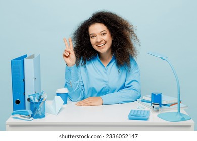 Young smiling cheerful successful employee business woman wear casual shirt sit work at white office desk look camera show v-sign gesture isolated on plain pastel light blue background studio portrait