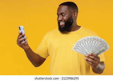 Young smiling cheerful winner happy black man 20s wear bright casual t-shirt holding fan of cash money in dollar banknotes mobile cell phone isolated on plain yellow color background studio portrait.