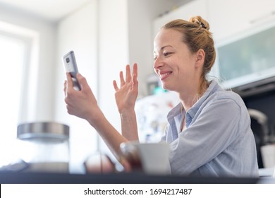 Young Smiling Cheerful Woman Indoors At Home Kitchen Using Social Media On Phone For Video Chatting And Stying Connected With Her Loved Ones. Stay At Home, Social Distancing Lifestyle.