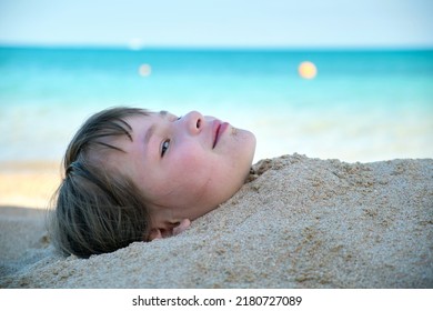 Young Smiling Child Girl Lying Down Covered With White Sand On Tropic Beach On Blue Sky And Ocean Water Background