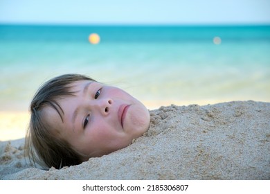 Young Smiling Child Girl Lying Down Covered With White Sand On Tropic Beach On Blue Sky And Ocean Water Background