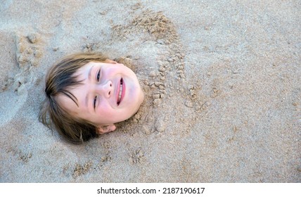 Young Smiling Child Girl Lying Down Covered With White Sand On Tropic Beach On Blue Sky And Ocean Water Background