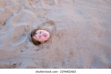 Young Smiling Child Girl Lying Down Covered With White Sand On Tropic Beach On Blue Sky And Ocean Water Background