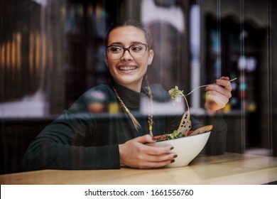 Young Smiling College Girl Sitting In Restaurant, Having Delicious Salad For Lunch And Looking Trough Window. She Is On The Break Between Classes. Picture Taken From Outside.