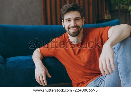 Young smiling cool bachelor man wears red t-shirt look camera sit on blue sofa couch stay at home hotel flat rest relax spend free spare time in living room indoors grey wall. People lounge concept