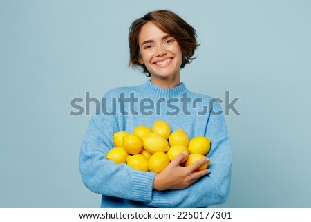 Young smiling cool positive healthy caucasian woman wear knitted sweater look camera hold pile of lemons isolated on plain pastel light blue cyan background studio portrait. People lifestyle concept