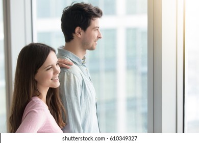 Young Smiling Couple Looking Through Window At Big City Scenery At Apartment Or Office, Pensive Man And Beautiful Woman Dreaming And Planning Future, Real Estate Purchasing, Renting Flat 