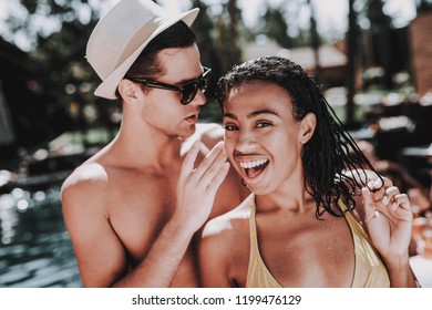 Young Smiling Couple Talking At Pool Party. Young Man Wearing Hat And Sunglasses Whispering To Beautiful Black Woman In Bikini At Poolside. Happy Friends Enoying Pool Party. Summer Vacation Concept