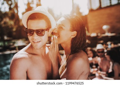 Young Smiling Couple Talking At Pool Party. Beautiful Black Woman In Bikini Whispering To Young Man Wearing Hat And Sunglasses At Poolside. Happy Friends Enoying Pool Party. Summer Vacation Concept