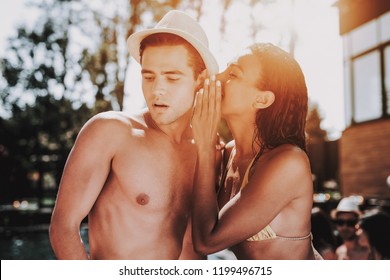 Young Smiling Couple Talking At Pool Party. Beautiful Black Woman In Bikini Whispering To Young Man Wearing Hat And Sunglasses At Poolside. Happy Friends Enoying Pool Party. Summer Vacation Concept