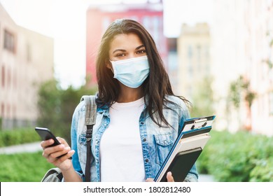 Young Smiling  Dark Skinned Girl Student Wearing  Medical Mask And Walking At The University Campus With Mobile Phone And Pile Of Books In Hands.