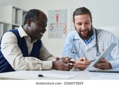 Young smiling doctor pointing at tablet screen with results of medical test and explaining diagnosis to senior African American male patient - Powered by Shutterstock