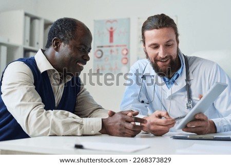 Young smiling doctor pointing at tablet screen with results of medical test and explaining diagnosis to senior African American male patient
