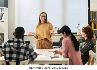 Young smiling educator explaining concepts to diverse group of students studying foreign languages in classroom setting 
