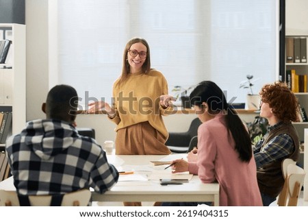 Young smiling educator explaining concepts to diverse group of students studying foreign languages in classroom setting 