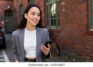 Young Smiling Elegant Asian Busy Business Woman Leader Executive Wearing Suit Standing Outside Office Using Cell Phone Applications. Smiling Professional Chinese Woman Holding Smartphone Outdoors.
