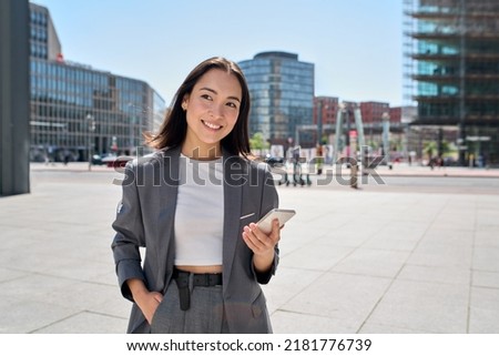 Young smiling elegant Asian busy business woman leader wearing suit standing in big city using cell phone platform applications. Smiling woman holding smartphone walking on street outdoors.