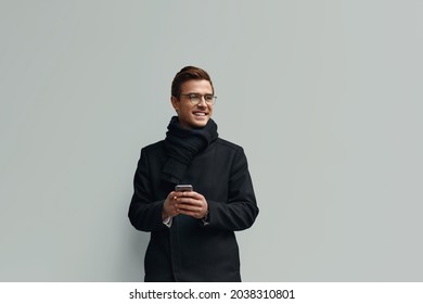 Young smiling european businessman standing and using mobile phone. Concept of modern successful man. Handsome stylish guy wearing scarf, coat and glasses. Isolated on white background. Copy space