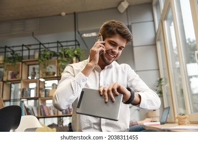Young smiling european businessman talking on mobile phone and watching time on wristwatch. Concept of modern successful man. Bearded stylish guy wearing formal clothes. Office interior. Daytime