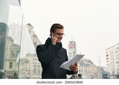 Young Smiling European Businessman Talking On Mobile Phone And Looking On Document Papers In City. Concept Of Modern Successful Man. Handsome Stylish Guy Wearing Scarf, Coat And Glasses. Daytime