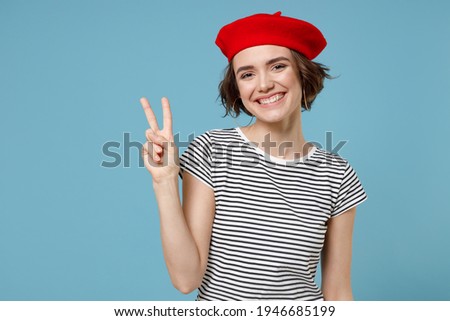 Young smiling european woman 20s with short hairdo wear french beret red hat striped t-shirt show victory v-sign gesture isolated on pastel blue background studio portrait. People lifestyle concept