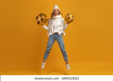 young smiling excited pretty woman jumping holding golden present boxes celebrating new year, christmas gifts, wearing white knitted sweater, scarf and hat, winter fashion, on yellow background
