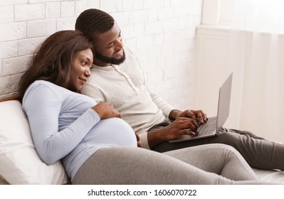 Young smiling expecting woman and her happy husband using laptop while relaxing on bed at home, checking on furniture for baby room, copy space