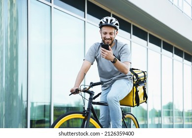 Young smiling express food delivery courier riding bicycle with insulated bag behind his back is looking at his phone.