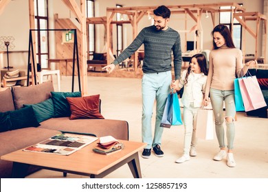 Young Smiling Family With Purchase Bags. Furniture Store Concept. Sunny Day. Good Mood. Loving Each Other. Bright Light. Happy Together. Going Shopping. Look At The Couch. Different Colors.