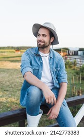 young smiling farmer in brim hat sitting on wooden fence on farmland